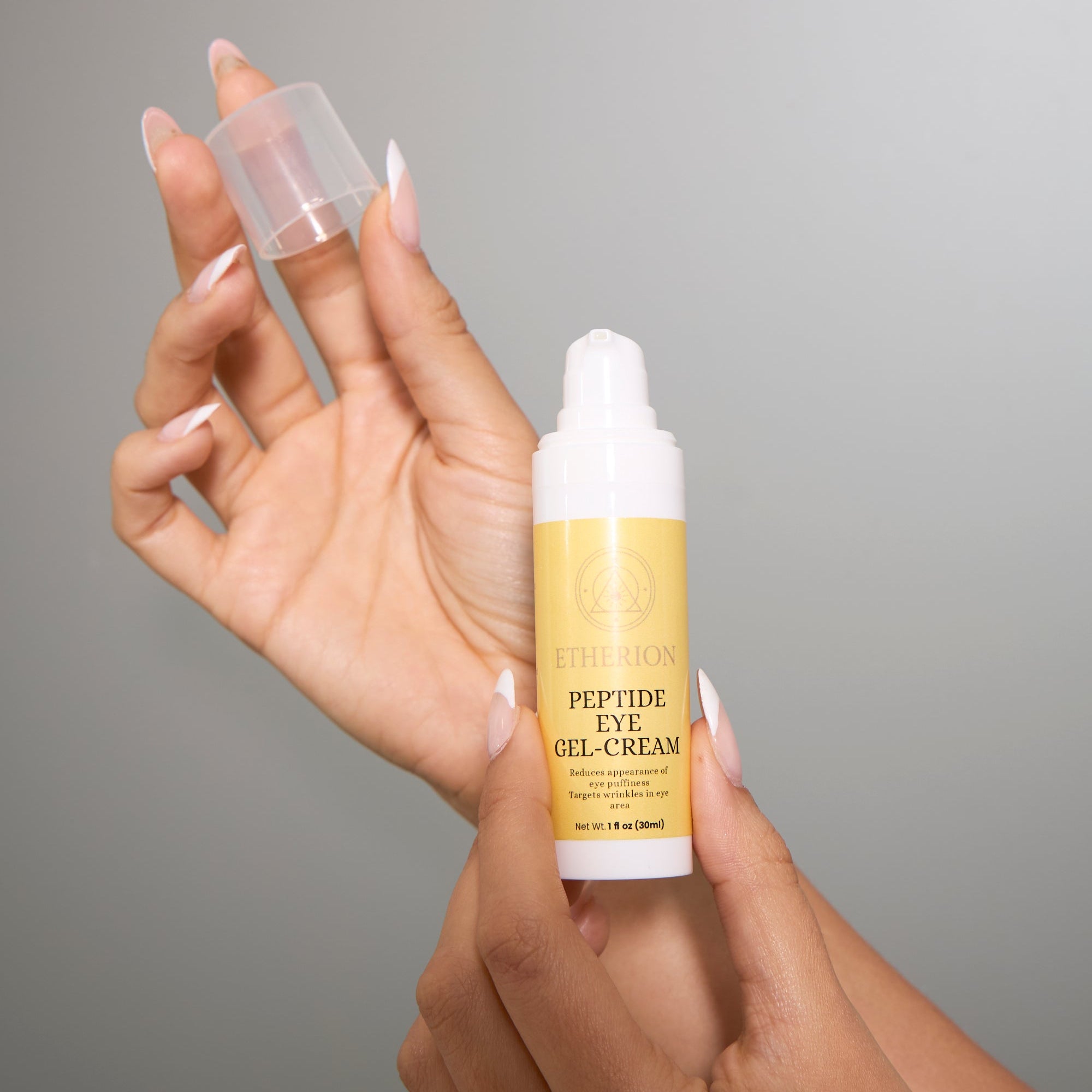 Hand holding a bottle of Etherion Peptide Eye Gel-Cream against a gray background